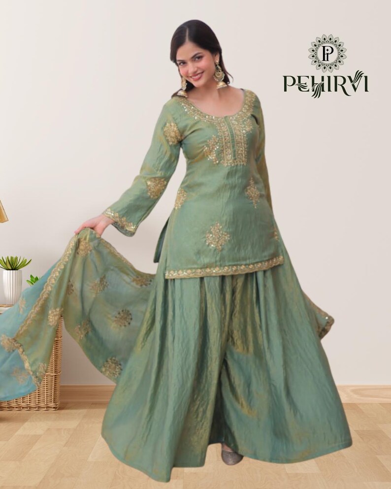 May include: A seafoam green Indian salwar kameez set with gold embroidery. The set includes a long-sleeved tunic, wide-legged pants, and a matching scarf. The tunic and pants are embellished with intricate gold detailing. The outfit is worn by a person.