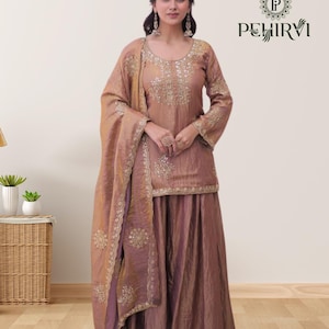 May include: A mauve-colored, embroidered salwar kameez set with a matching dupatta. The long-sleeved top and skirt are embellished with gold-colored embroidery. The dupatta is draped over the shoulder. The brand name "PEHIRVI" is visible in the upper right corner.
