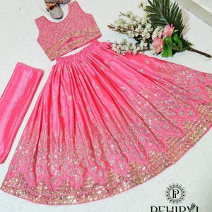 May include: A pink, embroidered three-piece outfit, including a sleeveless top, a long skirt, and a matching scarf. The skirt and top are embellished with gold-colored embroidery. A pair of small, embellished shoes and a bouquet of flowers are also present.