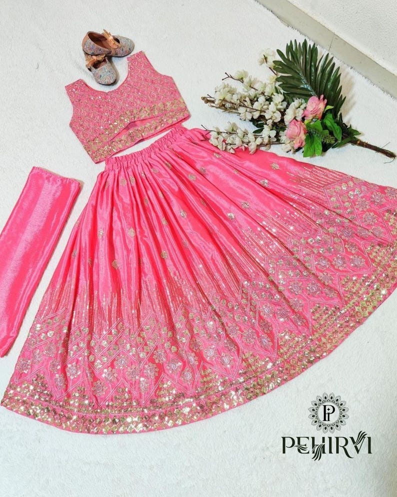 May include: A pink, embroidered three-piece outfit, including a sleeveless top, a long skirt, and a matching scarf. The skirt and top are embellished with gold-colored embroidery. A pair of small, embellished shoes and a bouquet of flowers are also present.