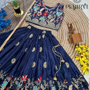 Op de afbeelding: Een marineblauwe tweedelige outfit met een crop top en een lange rok. De top en rok zijn versierd met gouden, rode en blauwe bloemenborduursels. De outfit wordt gepresenteerd op een geweven mat.