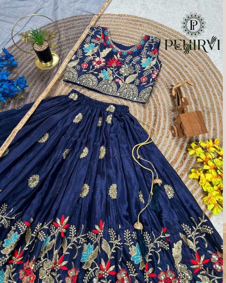 Op de afbeelding: Een marineblauwe tweedelige outfit met een crop top en een lange rok. De top en rok zijn versierd met gouden, rode en blauwe bloemenborduursels. De outfit wordt gepresenteerd op een geweven mat.