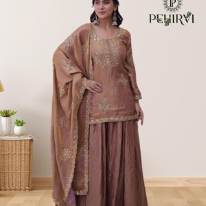 May include: A formal, two-piece outfit in a muted rose color, featuring intricate gold embroidery. The ensemble includes a long-sleeved top and a wide-legged skirt, complemented by a matching shawl. The outfit is from the brand PEHIRVI.