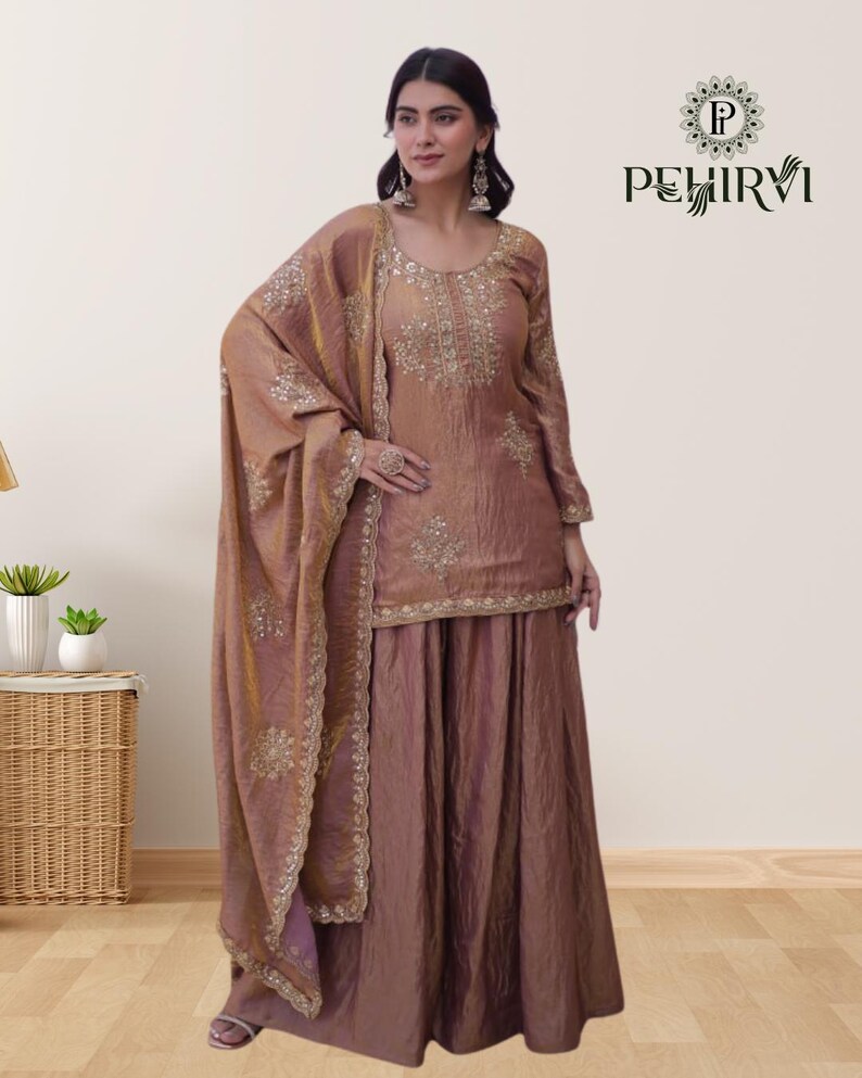 May include: A formal, two-piece outfit in a muted rose color, featuring intricate gold embroidery. The ensemble includes a long-sleeved top and a wide-legged skirt, complemented by a matching shawl. The outfit is from the brand PEHIRVI.