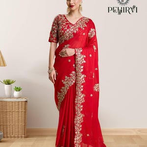 Puede incluir: Un sari rojo con bordado floral dorado. El sari presenta una blusa bordada a juego con mangas cortas y cuello en V. El sari está drapeado sobre el cuerpo, mostrando el intrincado diseño y la tela fluida.