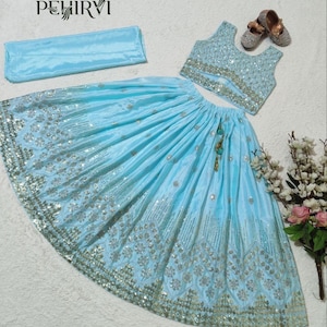 May include: A light blue, three-piece traditional outfit. The set includes a sleeveless top, a long skirt with silver embellishments, and a matching scarf. The skirt and top are adorned with intricate silver embroidery. The outfit is displayed with a pair of small, embellished shoes.