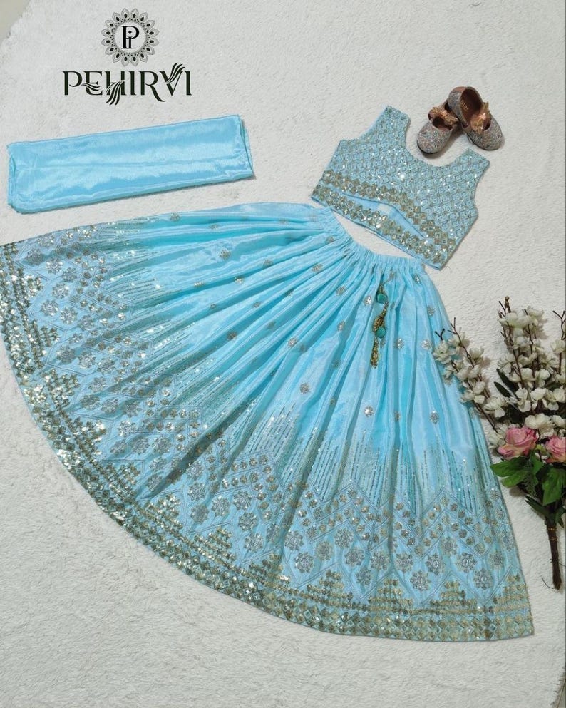 May include: A light blue, three-piece traditional outfit. The set includes a sleeveless top, a long skirt with silver embellishments, and a matching scarf. The skirt and top are adorned with intricate silver embroidery. The outfit is displayed with a pair of small, embellished shoes.