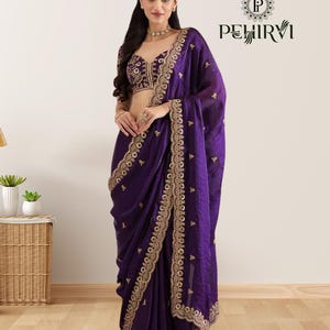 Puede incluir: Un sari de color púrpura intenso con intrincados bordados dorados a lo largo de los bordes y el corpiño. El sari está elegantemente drapeado, mostrando el diseño y la textura detallados. También se muestra una blusa a juego con adornos dorados.