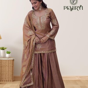 May include: A brown, three-piece ethnic outfit featuring a long-sleeved top, a wide skirt, and a matching scarf. The outfit is embellished with intricate gold embroidery around the neckline, cuffs, and hem. The brand name "PEHIRVI" is displayed in the upper right corner.