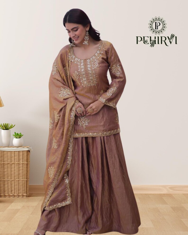 May include: A brown, three-piece ethnic outfit featuring a long-sleeved top, a wide skirt, and a matching scarf. The outfit is embellished with intricate gold embroidery around the neckline, cuffs, and hem. The brand name "PEHIRVI" is displayed in the upper right corner.