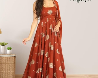 May include: A floor-length rust-colored sleeveless dress featuring a square neckline and a flared skirt adorned with a repeating gold floral pattern. The garment includes a matching rust-colored shawl draped over one shoulder. The text PEHIRVI is visible.