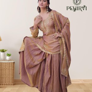 May include: A woman wearing a mauve-colored traditional Indian outfit. The ensemble includes a long-sleeved top with intricate gold embroidery, a matching skirt, and a draped shawl. The outfit is embellished with gold accents and the brand name "PEHIRVI" is visible.