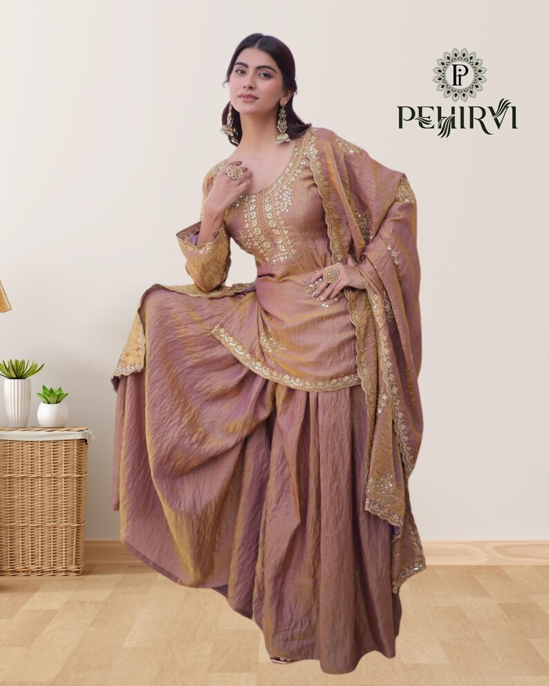 May include: A woman wearing a mauve-colored traditional Indian outfit. The ensemble includes a long-sleeved top with intricate gold embroidery, a matching skirt, and a draped shawl. The outfit is embellished with gold accents and the brand name "PEHIRVI" is visible.