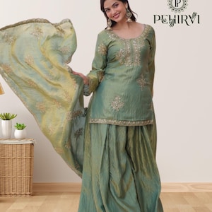 May include: A green silk salwar kameez set with intricate embroidery and embellishments. The set includes a long-sleeved tunic, wide-legged pants, and a matching dupatta. The outfit is worn by a person, and the dupatta is flowing in the air.