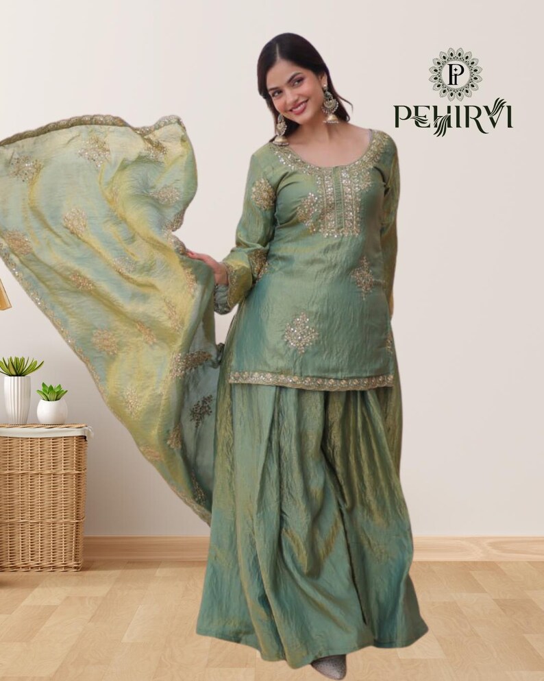 May include: A green silk salwar kameez set with intricate embroidery and embellishments. The set includes a long-sleeved tunic, wide-legged pants, and a matching dupatta. The outfit is worn by a person, and the dupatta is flowing in the air.