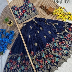 Op de afbeelding: Een marineblauwe tweedelige outfit met een crop top en een lange rok. De top en rok zijn versierd met ingewikkelde bloemenborduursels in rood, goud en teal. De outfit wordt gepresenteerd op een getextureerd oppervlak met decoratieve elementen.
