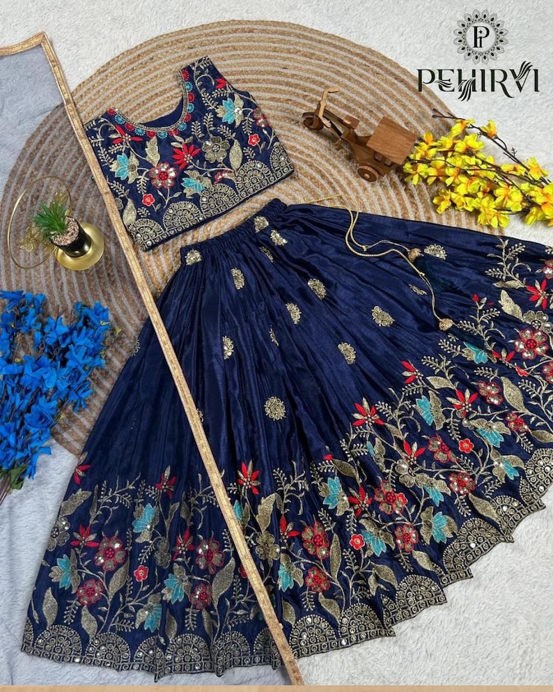 Op de afbeelding: Een marineblauwe tweedelige outfit met een crop top en een lange rok. De top en rok zijn versierd met ingewikkelde bloemenborduursels in rood, goud en teal. De outfit wordt gepresenteerd op een getextureerd oppervlak met decoratieve elementen.