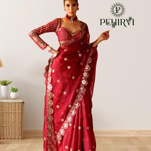 May include: A deep red saree with intricate gold and silver embellishments. The saree features a detailed border and a matching blouse with long sleeves. The design includes floral patterns and a decorative neckline. The saree is a traditional garment.
