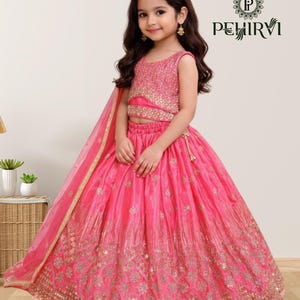 May include: A young girl wearing a vibrant pink lehenga choli, a traditional Indian outfit. The outfit features intricate gold embroidery and a matching dupatta. The top is sleeveless and embellished with sequins. The brand name "PEHIRVI" is visible.