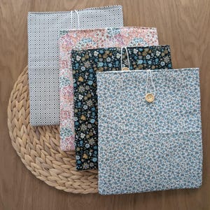 Puede incluir: Cuatro fundas de libros de tela con estampados florales y geométricos. Cada funda es rectangular y presenta un diseño diferente, incluyendo pequeñas flores azules sobre fondo blanco y un fondo negro con flores azules y amarillas. Se aseguran con un cordón y un botón.