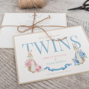 Puede incluir: Una tarjeta azul claro y blanca con el texto "Were Baptising our TWINS Amelia Suzanne & Noah Joshua" e ilustraciones de dos conejos, uno con una bufanda rosa y el otro con una bufanda azul.