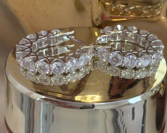 Puede incluir: Un par de pendientes de aro plateados, cada uno incrustado con múltiples piedras preciosas redondas y transparentes. Los pendientes se exhiben sobre una superficie reflectante de color dorado, creando un efecto brillante. Las piedras están espaciadas uniformemente.
