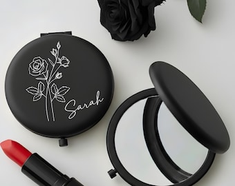 May include: Two black compact mirrors, one open, with a white floral design and the name "Sarah". A red lipstick and a black rose are also present. The mirrors are round and have a matte finish, ideal for a handbag.