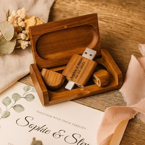 Könnte beinhalten: Ein hölzerner USB-Stick mit der Gravur "Sophie & Steve 10-25-2025" liegt in einer offenen Holzbox. Die Box enthält zwei Holzstücke und steht auf einem Tisch mit einer Hochzeitseinladung und floralen Akzenten. Die Farbpalette ist warm und natürlich.