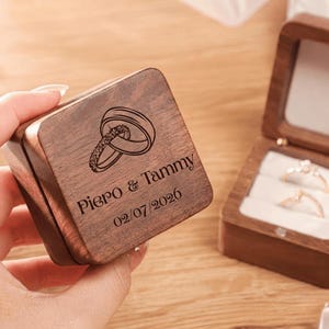 May include: A square dark wood ring box with a hinged lid featuring an engraved graphic of two interlocking rings, the names Piero & Tammy, and the date 02/07/2026. The open box interior contains white velvet inserts holding two rings.