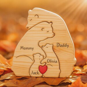 Puede incluir: Rompecabezas familiar de osos de madera con las palabras "Mommy", "Daddy", "Olivia" y "Jack" grabadas. Un coraz&oacute;n rojo est&aacute; en el centro. El rompecabezas es de madera clara y est&aacute; sobre un fondo borroso de hojas de oto&ntilde;o.