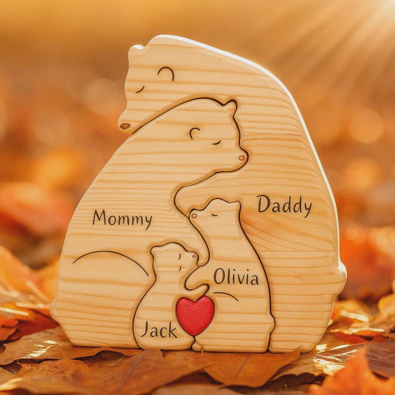 Puede incluir: Rompecabezas familiar de osos de madera con las palabras "Mommy", "Daddy", "Olivia" y "Jack" grabadas. Un coraz&oacute;n rojo est&aacute; en el centro. El rompecabezas es de madera clara y est&aacute; sobre un fondo borroso de hojas de oto&ntilde;o.