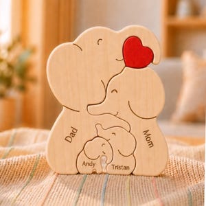 May include: A light-colored wooden puzzle shaped like a family of four elephants. The puzzle features a bright red heart at the top. The names Dad, Mom, Andy, and Tristan are engraved on the individual elephant-shaped wooden puzzle pieces.
