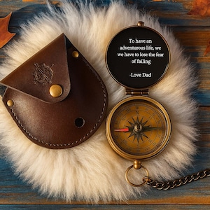 May include: A vintage brass compass with a leather case, open to reveal a quote: "To have an adventurous life, we have to lose the fear of failing -Love Dad." The compass has a detailed face and a chain. The case is brown.