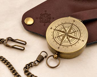 May include: A round brass compass with a detailed wind rose design on the lid, featuring cardinal directions N, S, E, and W. It is attached to a metallic chain and rests on a dark brown leather case with a stamped compass emblem and brass snap.
