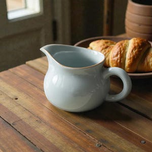 May include: A light blue ceramic pitcher with a rounded body and a curved handle. The pitcher has a gold rim and sits on a wooden table. A plate with croissants is in the background.