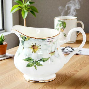 Könnte beinhalten: Weißer Keramik-Sahnegießer mit floralem Design mit großen weißen Blüten und grünen Blättern. Der Sahnegießer hat einen goldenen Rand und Griff. Eine passende Tasse ist im Hintergrund. Der Sahnegießer steht auf einer Holzoberfläche.