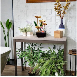 May include: A decorative indoor water fountain with gold-colored lotus flower accents sits on a gray table. The table also holds potted plants and a vase with gold-colored branches. The room has a white tile wall and a variety of green plants.