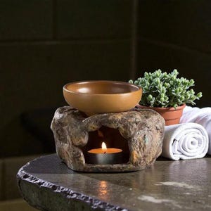 May include: A stone-effect essential oil diffuser with a brown ceramic bowl. A lit tea light candle is inside the diffuser. A small potted plant and rolled white towel are also present.