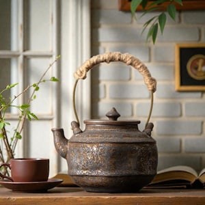 Puede incluir: Una tetera de cerámica marrón con un asa de cuerda y una taza y platillo a juego. La tetera tiene una superficie texturizada y una tapa pequeña y puntiaguda. El asa está hecha de una cuerda gruesa y tejida.