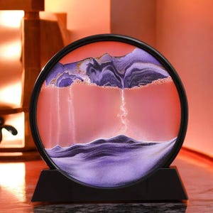 Peut inclure: Une image d'art de sable circulaire avec un cadre et un support noirs. Le paysage présente des montagnes violettes et dorées, un ciel rose et du sable blanc qui coule. La base est noire.