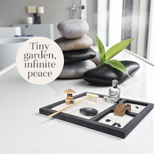 Pode incluir: Um conjunto de jardim zen com uma moldura preta, areia branca e acessórios em miniatura, incluindo um pagode de madeira, um ancinho e uma estátua de Buda prateada. A imagem também apresenta uma pilha de pedras e o texto "Tiny garden, infinite peace."
