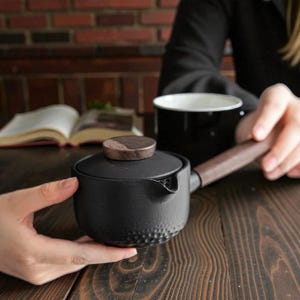 May include: A black ceramic teapot with a wooden handle and lid. The teapot has a textured lower half and a small spout. A black mug and an open book are in the background. The teapot is held by two hands.