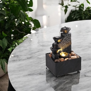 May include: A small, black tabletop fountain with water cascading down tiered bowls. The fountain has a textured, speckled finish and is illuminated with a warm light. It sits on a base filled with small stones, creating a tranquil ambiance.