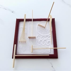 Puede incluir: Un pequeño jardín zen con un marco de madera rojo oscuro. La arena blanca está rastrillada con varias herramientas de madera, creando patrones. Las herramientas incluyen rastrillos y una pequeña pala. El jardín es de forma cuadrada.