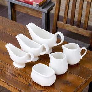 May include: A selection of white ceramic serving dishes, including gravy boats and small jugs, displayed on a wooden table. The gravy boats have handles and spouts, while the jugs have curved handles. The pieces are clean and simple.