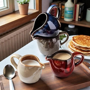 May include: A stack of four ceramic cream pitchers in various colors: navy blue, brown, beige, and red. Each pitcher has a handle and a spout. One beige pitcher is filled with milk, and one red pitcher is filled with a brown liquid.
