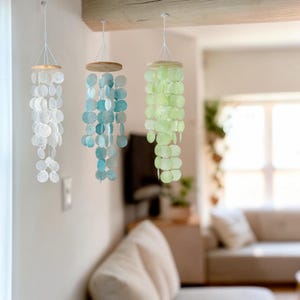 May include: Three hanging wind chimes with circular, iridescent shell pieces in white, blue, and green. Each chime has a wooden top and is suspended by white string. The chimes are hanging indoors, adding a touch of coastal decor.