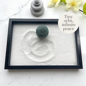 Może przedstawiać: Miniaturowy ogród zen z czarną ramą, białym piaskiem i turkusową kulą ze spiralnym wzorem. Piasek jest zagrabiony okrągłym wzorem. Widoczny jest również mały stos szarych kamieni i tekst "Tiny orbs, infinite peace".