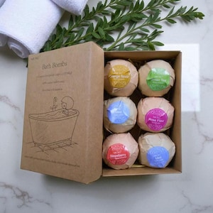 May include: A box of bath bombs with various flavors, including orange, green tea, rose, strawberry, and mint. The box is brown with the words "Bath Bombs" and a drawing of a person in a bathtub. White towels and greenery are in the background.