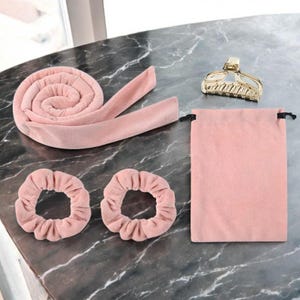 Puede incluir: Un juego de rizado de cabello rosa que incluye una pieza larga de tela enrollada, dos coleteros, una bolsa con cordón y una pinza para el cabello dorada. El juego está diseñado para crear rizos sin calor y se muestra sobre una superficie con vetas de mármol.